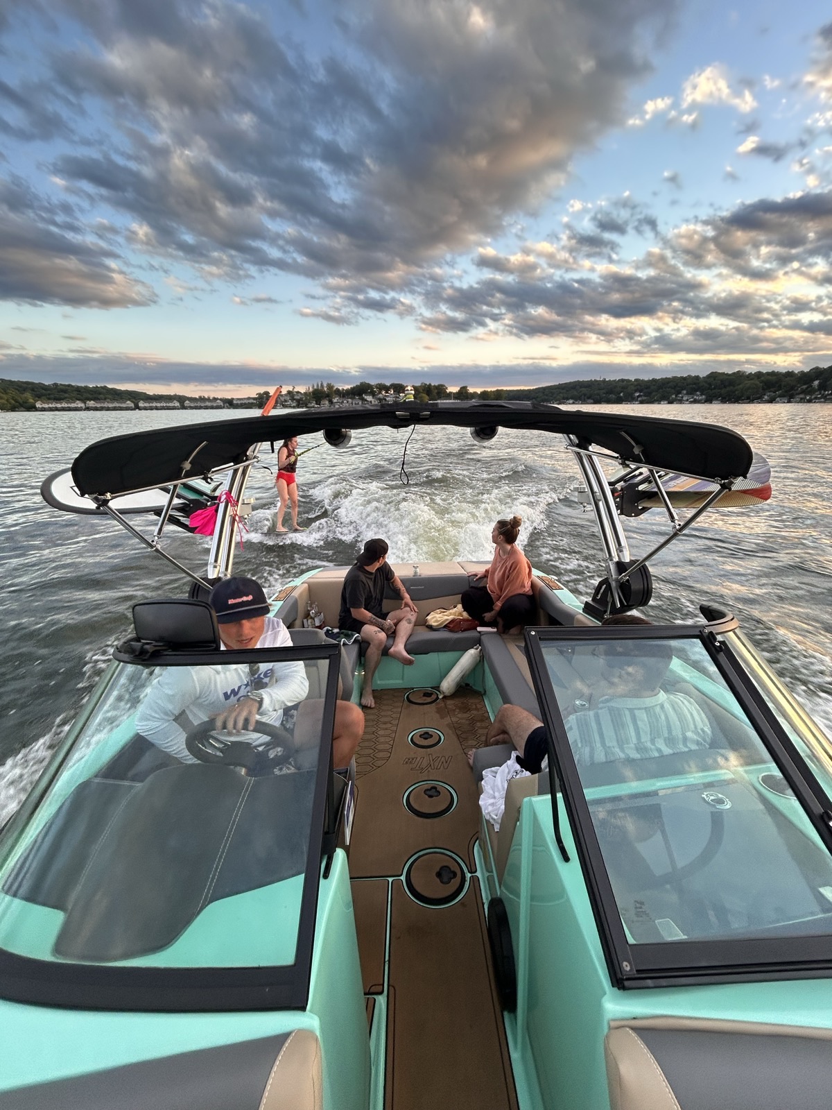 A family relaxing on the boat at sunset while a rider surfs the wake behind on Lake Hopatcong